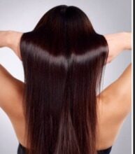 एक खुश महिला अपने लहराते, चमकदार, सिल्की-स्मूथ बालों को छू रही है। बैकग्राउंड में स्वस्थ बालों के लिए नारियल तेल, दही और एलोवेरा जैसे प्राकृतिक उपचार सामग्री रखी हैं।