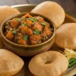 एक पीतल की थाली में गरमा गरम, सुनहरे रंग की फूली हुई पूरियों का ढेर, जिसके बगल में एक कटोरी में खट्टी-मीठी और मसालेदार कद्दू की सब्ज़ी (सीताफल) रखी है। सब्ज़ी पर हरा धनिया गार्निश किया गया है।