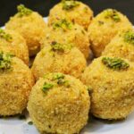 सुनहरी भूरे रंग के, मेवों से सजे हुए पारंपरिक मूंग दाल के लड्डू, एक सादे प्लेट में रखे हुए हैं।