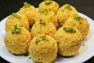 सुनहरी भूरे रंग के, मेवों से सजे हुए पारंपरिक मूंग दाल के लड्डू, एक सादे प्लेट में रखे हुए हैं।