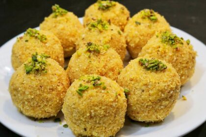 सुनहरी भूरे रंग के, मेवों से सजे हुए पारंपरिक मूंग दाल के लड्डू, एक सादे प्लेट में रखे हुए हैं।