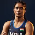 भारतीय महिला पहलवान विनेश फोगाट मैट पर आत्मविश्वास से भरी मुद्रा में खड़ी हैं, सामने 2028 लॉस एंजिल्स ओलंपिक का लोगो लगा है।