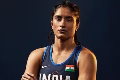 भारतीय महिला पहलवान विनेश फोगाट मैट पर आत्मविश्वास से भरी मुद्रा में खड़ी हैं, सामने 2028 लॉस एंजिल्स ओलंपिक का लोगो लगा है।