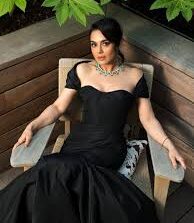 ब्लैक ड्रेस पहने एक अभिनेत्री, आउटडोर लकड़ी की सीट पर आत्मविश्वास के साथ बैठी हुई