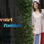 एक खूबसूरत साउथ एशियन महिला पारंपरिक घर के दरवाजे पर खड़ी है, चारों तरफ हरियाली और फूल हैं। उसने मल्टीकलर प्रिंटेड कुर्ता, स्कर्ट और दुपट्टा पहना हुआ है। बाईं ओर “Navratri Fashion” लिखा हुआ है।