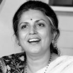 काले-सफेद (Black & White) पोर्ट्रेट में एक महिला मुस्कुराते हुए दिखाई दे रही हैं। उन्होंने साड़ी पहन रखी है, माथे पर बिंदी है और बाल पीछे की ओर बंधे हुए हैं। तस्वीर में उनका चेहरा आत्मविश्वास, सादगी और सौम्यता को दर्शाता है। बैकग्राउंड में घर का इंटीरियर दिखाई दे रहा है, जिससे यह एक अनौपचारिक और स्वाभाविक क्षण की तस्वीर लगती है।
