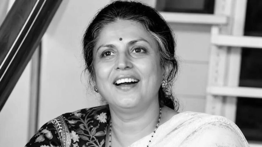 काले-सफेद (Black & White) पोर्ट्रेट में एक महिला मुस्कुराते हुए दिखाई दे रही हैं। उन्होंने साड़ी पहन रखी है, माथे पर बिंदी है और बाल पीछे की ओर बंधे हुए हैं। तस्वीर में उनका चेहरा आत्मविश्वास, सादगी और सौम्यता को दर्शाता है। बैकग्राउंड में घर का इंटीरियर दिखाई दे रहा है, जिससे यह एक अनौपचारिक और स्वाभाविक क्षण की तस्वीर लगती है।