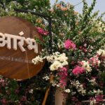 फूलों से सजे प्रवेश द्वार पर लगा आरंभ (Aarambh) कैफे का लकड़ी का साइनबोर्ड, जो सात्विक और मिलेट आधारित भोजन की पहचान दर्शाता है।