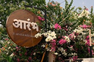 फूलों से सजे प्रवेश द्वार पर लगा आरंभ (Aarambh) कैफे का लकड़ी का साइनबोर्ड, जो सात्विक और मिलेट आधारित भोजन की पहचान दर्शाता है।