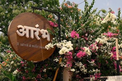 फूलों से सजे प्रवेश द्वार पर लगा आरंभ (Aarambh) कैफे का लकड़ी का साइनबोर्ड, जो सात्विक और मिलेट आधारित भोजन की पहचान दर्शाता है।