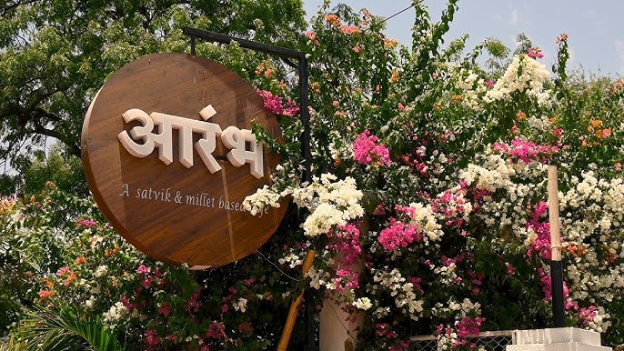 फूलों से सजे प्रवेश द्वार पर लगा आरंभ (Aarambh) कैफे का लकड़ी का साइनबोर्ड, जो सात्विक और मिलेट आधारित भोजन की पहचान दर्शाता है।