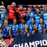 भारतीय महिला हॉकी टीम FIH Women’s Nations Cup जीतने के बाद ट्रॉफी और पदकों के साथ मंच पर जश्न मनाती हुई दिखाई दे रही है। खिलाड़ी खुशी से उत्साहित होकर "Champions" बोर्ड के सामने जीत का उत्सव मना रही हैं।
