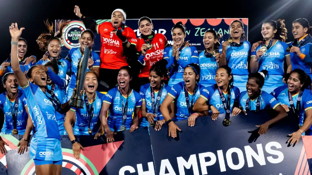 भारतीय महिला हॉकी टीम FIH Women’s Nations Cup जीतने के बाद ट्रॉफी और पदकों के साथ मंच पर जश्न मनाती हुई दिखाई दे रही है। खिलाड़ी खुशी से उत्साहित होकर "Champions" बोर्ड के सामने जीत का उत्सव मना रही हैं।