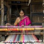 An Indian woman weaving a colorful silk fabric on a traditional handloom inside a rural weaving workshop.