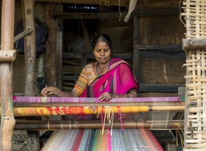 An Indian woman weaving a colorful silk fabric on a traditional handloom inside a rural weaving workshop.