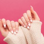 Hands showcasing stylish nude nails with white heart nail art against a pink background.