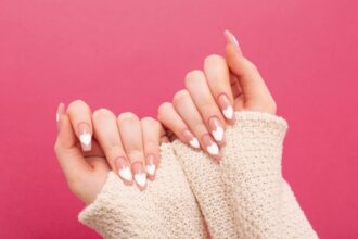 Hands showcasing stylish nude nails with white heart nail art against a pink background.