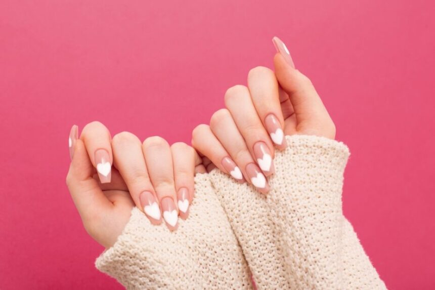 Hands showcasing stylish nude nails with white heart nail art against a pink background.